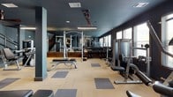 This two-story fitness center combines functional design with a sophisticated aesthetic, characterized by deep teal-painted walls, neutral carpet tile flooring, and linear pendant lighting throughout. The main workout floor is thoughtfully organized into dedicated zones, featuring a row of treadmills and ellipticals positioned along large windows that allow natural light to filter in. Full-length mirrors amplify the sense of space and aid in monitoring form during exercise. A comprehensive free weight section includes a fully stocked dumbbell rack, adjustable benches, and a power rack, while a separate area houses an array of selectorized strength machines suited for full-body training. Ceiling fans provide additional air circulation across the workout floor. A service alcove offers cubbies stocked with towels and sundry items alongside frosted-glass doors leading to additional rooms. The entrance corridor features a wood-paneled accent wall displaying Western-inspired artwork, adding a distinctive character to the space. The second floor, accessible via a staircase with a sleek cable-rail system, provides additional open workout space illuminated by wall sconces and pendant lighting, completing this well-appointed fitness facility.