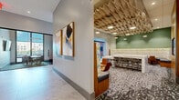 Located at 12 Church Street in New Rochelle, NY, this single-floor office suite offers a refined and contemporary workspace with a well-considered layout. A wide, light-filled hallway with large-format porcelain tile flooring and curated abstract artwork sets an elegant tone upon entry, guiding visitors toward the suite's various spaces.

The centerpiece of the suite is a striking collaborative lounge area, distinguished by a sculptural geometric wood ceiling installation adorned with pendant lighting. A substantial marble-topped communal table with bar-height stools anchors the space, complemented by upholstered banquette seating set against a sage green accent wall with a bold textural wallcovering. Patterned carpet tiles add visual depth to the floor throughout.

A built-in media nook features a wall-mounted display screen framed by rich wood paneling and set against a textured stone wall, creating an ideal space for presentations or informal gatherings. Three private offices provide enclosed workspace, one of which is enclosed by full-height glass walls, allowing natural light and urban views of New Rochelle to permeate the interior. The overall design balances professionalism with creativity, making this suite well-suited for a modern business environment.