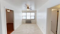 This single-floor space offers a clean, well-maintained interior defined by its bright and airy atmosphere. The main living area is finished with light-toned tile flooring that extends throughout, lending a crisp and cohesive look to the space. White walls provide a neutral backdrop, enhancing the sense of openness and allowing for versatile decorating possibilities. A multi-blade ceiling fan with an integrated light fixture serves as both a functional and understated focal point overhead. Natural light filters in through a set of windows positioned along one wall, brightening the room throughout the day. Beneath the windows sits a classic cast-iron radiator, reflecting the space's traditional heating infrastructure. The layout is straightforward and efficient, with an open flow that makes the interior feel welcoming. A doorway visible along one wall provides access to additional areas of the space, suggesting a connected and practical floor plan. Overall, this space combines simple, timeless finishes with functional features to create a comfortable and livable environment.