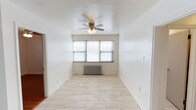 This single-floor space offers a clean, well-maintained interior defined by its bright and airy atmosphere. The main living area is finished with light-toned tile flooring that extends throughout, lending a crisp and cohesive look to the space. White walls provide a neutral backdrop, enhancing the sense of openness and allowing for versatile decorating possibilities. A multi-blade ceiling fan with an integrated light fixture serves as both a functional and understated focal point overhead. Natural light filters in through a set of windows positioned along one wall, brightening the room throughout the day. Beneath the windows sits a classic cast-iron radiator, reflecting the space's traditional heating infrastructure. The layout is straightforward and efficient, with an open flow that makes the interior feel welcoming. A doorway visible along one wall provides access to additional areas of the space, suggesting a connected and practical floor plan. Overall, this space combines simple, timeless finishes with functional features to create a comfortable and livable environment.
