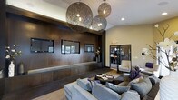 This single-floor space showcases a thoughtfully designed interior that blends modern aesthetics with a warm, inviting ambiance. The focal point of the main lounge area is a dramatic cluster of oversized woven sphere pendant lights that hang at varying heights from the ceiling, casting a soft, ambient glow throughout the room. The ceiling itself is finished in crisp white, providing contrast against the rich, dark wood wall paneling that anchors the space.

The feature wall is clad in deep espresso-toned wood and incorporates a built-in ledge with recessed window openings, adding architectural depth and visual interest. Dark hardwood flooring runs throughout, complementing the warm tones of the wall treatment and contributing to a cohesive, sophisticated palette.

The seating area is furnished with generously sized gray upholstered sofas arranged around a low, round coffee table, creating a comfortable gathering space. To one side, glass-paneled doors with dark frames lead into an adjacent room, where a tall chair and bar-height counter are visible, suggesting a secondary lounge or casual workspace. Recessed lighting supplements the pendant fixtures, ensuring the space is well-lit and functional throughout.