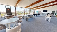 Located on Meadowlark Lane SE in Rio Rancho, New Mexico, this generously sized single-floor recreation and gathering space offers a well-appointed, open-plan layout designed for both leisure and social interaction. The room's most striking architectural feature is its vaulted ceiling lined with rich, exposed wood beams that run the length of the space, adding warmth and character to the otherwise contemporary interior.

Floor-to-ceiling windows along one full wall flood the interior with abundant natural light while providing views of the surrounding Rio Rancho streetscape. The blue-grey carpet tiles provide a neutral, cohesive foundation throughout. Globe pendant lights hang from the beamed ceiling, complementing the modern aesthetic.

The space is thoughtfully divided into distinct activity zones. A games area includes a pool table, foosball table, shuffleboard, and a raised cornhole platform positioned directly in front of the expansive windows. A lounge and dining area features round tables with upholstered chairs, benches, and a bar-height counter with stools beneath a wall-mounted flat-screen TV. Decorative artwork and potted plants add visual interest throughout.

A separate anteroom with soft grey walls, carpeted flooring, and glass-paned French doors provides a transitional entry point into the main recreation area, completing this versatile and inviting community space.