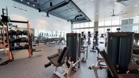 This single-floor fitness center offers a thoughtfully designed workout environment that combines functionality with an open, airy atmosphere. The space is defined by its impressively high ceilings, from which exposed ductwork and industrial-style track lighting extend, lending the facility a contemporary, urban aesthetic.

Large floor-to-ceiling windows line the perimeter, allowing abundant natural light to fill the interior and providing views of the outdoors, creating an energizing environment for exercise. Full-length mirrors are positioned along one wall, enhancing the sense of openness and supporting proper form during workouts.

The facility is comprehensively equipped with a diverse array of strength-training machines, including cable systems and resistance equipment, arranged to allow comfortable circulation throughout the space. A dedicated free weights area features an organized rack of medicine balls and dumbbells, catering to a variety of training styles. Toward the rear, a row of cardio machines—including treadmills and ellipticals—offers ample options for cardiovascular training.

The flooring combines durable carpet and hard surface zones suited to different workout activities. The overall layout is spacious and uncluttered, ensuring a comfortable and professional fitness experience. The industrial-modern design elements and generous natural light make this a standout workout facility.