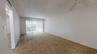 This single-story residence offers a thoughtfully laid-out floor plan visible across its multiple rooms. The living area is generously proportioned, featuring warm beige carpeting, white walls, and a textured ceiling with a ceiling fan. A large sliding glass door fitted with vertical blinds allows abundant natural light to fill the space while providing access to an outdoor area with greenery visible beyond.

The kitchen is efficiently designed in a galley configuration, featuring white cabinetry, granite-style countertops, wood-look flooring, and white appliances including a refrigerator, electric range, and under-cabinet range hood. A window above the sink brings in additional natural light.

The bedrooms are carpeted and bright, each featuring windows with blinds. One bedroom includes a double closet with sliding doors, while another offers built-in shelving alongside a closet, providing versatile storage solutions. An additional room features louvered closet doors for further storage.

The bathroom is finished with white square tile wainscoting, a granite-style vanity countertop, a tub-shower combination, and wood-look flooring, presenting a clean and functional design throughout.