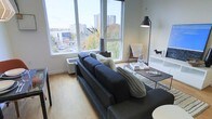 This contemporary single-floor residence is designed to maximize natural light and urban connectivity. The living area serves as the centerpiece of the space, anchored by large, floor-to-ceiling windows that frame expansive views of the surrounding cityscape, including treetop canopies and mid-rise buildings in the distance — an ever-changing backdrop through every season. Light streams generously into the room, creating a bright and welcoming atmosphere throughout the day.

The living space features light-toned hardwood flooring that complements the clean white walls and modern trim. A sleek, low-profile media console with a wall-mounted flat-screen television anchors one side of the room, while a stylish seating arrangement — centered on a dark upholstered sofa with layered textural cushions — defines the lounge area. A minimalist glass and metal coffee table adds to the contemporary aesthetic.

Thoughtful design details, including a bold floor lamp, a mid-century accent chair, and carefully curated décor, contribute to the space's modern yet inviting character. The open layout and generous window placement create a seamless connection between the interior and the vibrant urban environment outside, making this a well-appointed and visually engaging space.