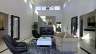 This single-floor space makes a striking impression through its contemporary architectural design and thoughtfully curated interiors. The main living area commands attention with dramatically high ceilings that amplify the sense of openness, while a generous arrangement of clerestory windows positioned near the roofline floods the space with natural light throughout the day. A bold, modern fireplace serves as the focal centerpiece of the room, framed by sleek tile surroundings and accented with decorative elements, including a vivid orange vase that adds a pop of color against the neutral palette. The furnishings are carefully selected to complement the modern aesthetic — a plush sectional sofa, a sculptural black leather accent chair, and a clean-lined white coffee table rest atop a textured area rug, creating a comfortable yet refined seating arrangement. The predominantly white walls and light-toned flooring enhance the bright, airy atmosphere, while dark wood and matte accents provide contrast and visual depth. A large ceiling fan adds both a functional and architectural touch overhead. The overall ambiance is one of modern elegance — open, luminous, and meticulously designed for both style and comfort.