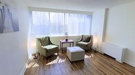 This single-floor space offers a welcoming and well-lit interior defined by its clean, modern design. The sitting area is a standout feature, anchored by a large picture window dressed with vertical blinds that diffuse abundant natural light throughout the room, casting soft patterns across the rich hardwood-style flooring. The window also frames views of lush greenery outside, adding a tranquil, nature-connected quality to the interior atmosphere.

The seating arrangement is thoughtfully composed, featuring two upholstered wingback-style chairs in a muted olive-green tone, positioned symmetrically on either side of a compact wooden side table. A tufted white ottoman at the center serves as both a functional footrest and a subtle focal point. Floor lamps on either side of the seating area provide warm supplemental lighting, enhancing the room's inviting ambiance during lower-light hours.

The flooring throughout features wood-look planks in warm, varied tones, contributing texture and warmth to the otherwise light and neutral color palette. White walls and bright natural light give the space an open, airy feel, making it both functional and visually comfortable.
