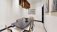Located at 1625 Northwest 20th Street in Miami, FL, this dedicated dining room is a well-appointed space that combines modern elegance with tropical flair. The room is anchored by a striking dark espresso-toned rectangular dining table, surrounded by a set of cane-back chairs that blend natural texture with contemporary design. The most dramatic architectural feature is the statement chandelier overhead — a sculptural piece crafted from layered bronze-toned slats that draw the eye upward and reinforce the room's elevated ceiling height, contributing to an airy, open ambiance. The crisp white walls serve as a clean canvas for a curated gallery of large-scale botanical artwork, depicting monochromatic palm fronds in warm wood frames. These pieces echo Miami's tropical character while maintaining a sophisticated, refined aesthetic. Black door frames and baseboards provide sharp contrast against the light-toned flooring, adding definition and a modern edge throughout the space. The overall design palette — a balance of dark wood, warm gold, natural cane, and white — creates an atmosphere that is both inviting and stylishly composed, making this dining room an ideal setting for intimate gatherings or formal dining experiences.