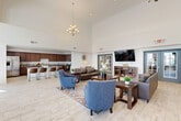 Located at 430 Mosby Blvd in Berryville, Virginia, this well-appointed single-floor community space offers a generously sized layout designed for both gathering and relaxation. The centerpiece is a dramatic open lounge area distinguished by soaring vaulted ceilings and an elegant chandelier, creating an airy, welcoming atmosphere. Plush leather sofas, accent chairs, and patterned area rugs define comfortable seating arrangements, while large windows along the perimeter flood the space with natural light and provide views of the surrounding exterior. A reading nook with open bookshelves and upholstered seating offers a quieter retreat within the lounge. The fully equipped kitchen features rich dark-wood cabinetry, granite countertops, stainless steel appliances including a range and refrigerator, and ample storage — ideal for communal events and catering. Light wood-look tile flooring runs throughout the kitchen and common areas, lending a clean, modern aesthetic. A secondary multipurpose room with light hardwood flooring, recessed lighting, and a wall-mounted television provides a versatile space suitable for presentations, screenings, or additional seating. Throughout the space, a neutral white and warm wood palette creates a cohesive, inviting environment suited for a variety of community activities.