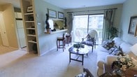 This single-floor residence offers a thoughtfully arranged living area that balances functionality with a warm, inviting ambiance. The space is anchored by plush carpeting in neutral tones, which complements the soft sage-green walls and creates a comfortable, cohesive atmosphere throughout.

The living area is well-lit, benefiting from a large sliding glass door that leads to a private balcony, allowing natural light to fill the room. The balcony provides views of the surrounding building exterior and outdoor surroundings, making it a pleasant extension of the interior living space.

A defining feature is the built-in open shelving unit along one wall, offering generous display and storage space for books and decorative items. Adjacent to this is a raised breakfast bar with barstool seating, cleverly separating the living area from a secondary functional zone while maintaining an open feel. Framed artwork above the bar adds a decorative touch.

The seating arrangement includes classic upholstered chairs and a sofa paired with a dark wood coffee table, creating an intimate and elegant gathering space. Decorative greenery and potted topiaries are placed throughout, lending a refined, traditional character to the overall design.