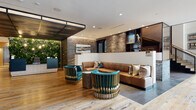 This thoughtfully designed single-floor space presents a sophisticated blend of industrial and contemporary aesthetics throughout its expansive lobby and lounge areas. Reclaimed hardwood flooring flows continuously throughout, anchoring each distinct zone while lending warmth and character to the overall environment.

The main entry features textured concrete-finish walls, a striking dark-tiled arched passageway, and glass-paneled entry doors that invite natural light inward. A dedicated reception area is positioned adjacent to a dramatic living green wall, framed by a dark steel ceiling detail and accented with Edison-style string lighting, creating an inviting, biophilic focal point.

A lounge seating area showcases tufted caramel leather banquette seating alongside bold teal-and-gold barrel chairs, with built-in display shelving set into exposed brick walls above. Curated artwork punctuates the space throughout.

A bright, window-lined lounge area features floor-to-ceiling glazing offering urban views, complemented by a warm wood ceiling panel, wingback chairs, velvet ottomans, and eclectic accent seating. An open staircase with industrial metal railings provides visual drama alongside double-height windows. The elevator alcove is finished with tactile cream tile walls, stainless steel doors, and a framed art piece. Together, these elements create a layered, design-forward environment that balances comfort with visual sophistication.