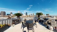 This rooftop deck in the heart of New Orleans, LA offers a dynamic combination of indoor and outdoor spaces designed for gathering and relaxation. The indoor lounge area makes an immediate impression with its bold color palette — vivid yellow upper walls contrast against cool teal-blue lower walls — and polished concrete floors that give the space an urban, modern edge. Comfortable seating with orange-cushioned chairs and a plush sofa is arranged around a coffee table, accompanied by vibrant artwork lining the walls. Recessed lighting and wall sconces provide a warm ambiance throughout. An adjacent indoor dining area features a large high-top table with ample seating, bathed in natural light streaming through expansive sliding glass doors that open directly onto the outdoor terrace. The open-air deck is the true centerpiece of this space, offering unobstructed panoramic views of the New Orleans skyline. Flanked by mature palm trees and enclosed by a white railing, the deck is furnished with a round dining set, lounge chairs, and a grill — perfect for outdoor entertaining. The seamless indoor-outdoor flow, combined with the breathtaking cityscape backdrop, makes this rooftop an exceptional venue above the vibrant streets of New Orleans.