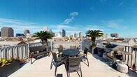 This rooftop deck in the heart of New Orleans, LA offers a dynamic combination of indoor and outdoor spaces designed for gathering and relaxation. The indoor lounge area makes an immediate impression with its bold color palette — vivid yellow upper walls contrast against cool teal-blue lower walls — and polished concrete floors that give the space an urban, modern edge. Comfortable seating with orange-cushioned chairs and a plush sofa is arranged around a coffee table, accompanied by vibrant artwork lining the walls. Recessed lighting and wall sconces provide a warm ambiance throughout. An adjacent indoor dining area features a large high-top table with ample seating, bathed in natural light streaming through expansive sliding glass doors that open directly onto the outdoor terrace. The open-air deck is the true centerpiece of this space, offering unobstructed panoramic views of the New Orleans skyline. Flanked by mature palm trees and enclosed by a white railing, the deck is furnished with a round dining set, lounge chairs, and a grill — perfect for outdoor entertaining. The seamless indoor-outdoor flow, combined with the breathtaking cityscape backdrop, makes this rooftop an exceptional venue above the vibrant streets of New Orleans.
