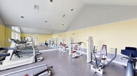 Located at 70 Greenway Square in Dover, Delaware, this fitness center offers a bright and functional workout environment designed to accommodate a range of exercise needs. The space is defined by its striking vaulted ceiling, which rises prominently and lends an open, airy feel throughout the room. Soft yellow walls, accented by a gray wainscoting band, create a clean and energizing atmosphere, while durable speckled flooring provides a practical surface suited for fitness activities.

Natural light flows generously into the space through large arched windows along one wall, offering views of the surrounding community and making the room feel welcoming and expansive. French doors provide an additional access point and further enhance the connection to the outdoors. A full-wall mirror runs along one side of the room, visually doubling the sense of space while allowing users to monitor their form during workouts.

The fitness center is well-equipped with a variety of cardio machines, including multiple treadmills and an elliptical trainer, as well as a selection of strength training machines with cushioned seats and weight stacks. Recessed lighting in the vaulted ceiling supplements the abundant natural light, ensuring the space remains well-lit at all times. Overall, this fitness center combines thoughtful architectural design with practical amenities to create an inviting and fully functional workout facility.