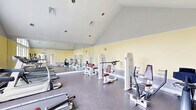 Located at 70 Greenway Square in Dover, Delaware, this fitness center offers a bright and functional workout environment designed to accommodate a range of exercise needs. The space is defined by its striking vaulted ceiling, which rises prominently and lends an open, airy feel throughout the room. Soft yellow walls, accented by a gray wainscoting band, create a clean and energizing atmosphere, while durable speckled flooring provides a practical surface suited for fitness activities.

Natural light flows generously into the space through large arched windows along one wall, offering views of the surrounding community and making the room feel welcoming and expansive. French doors provide an additional access point and further enhance the connection to the outdoors. A full-wall mirror runs along one side of the room, visually doubling the sense of space while allowing users to monitor their form during workouts.

The fitness center is well-equipped with a variety of cardio machines, including multiple treadmills and an elliptical trainer, as well as a selection of strength training machines with cushioned seats and weight stacks. Recessed lighting in the vaulted ceiling supplements the abundant natural light, ensuring the space remains well-lit at all times. Overall, this fitness center combines thoughtful architectural design with practical amenities to create an inviting and fully functional workout facility.