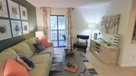 This single-floor residence at 6200 Dansey Dr in Raleigh, North Carolina, offers a well-appointed living space designed for comfortable everyday living. The living room serves as the heart of the home, showcasing a warm and inviting layout with neutral-toned carpeting that complements the overall color palette. A sliding glass door floods the room with natural light and provides direct access to a private balcony, where views of surrounding greenery can be enjoyed. Decorative floral curtains frame the door, adding a soft, stylish touch to the space. The living area comfortably accommodates a sectional sofa, an accent chair, and a central coffee table, creating a functional and aesthetically cohesive seating arrangement. A wooden media console along one wall offers ample storage and display space. One wall is painted in a muted olive green, providing a subtle contrast against the lighter surrounding walls and contributing to the room's warm ambiance. Recessed and floor lighting enhance the space during evening hours, ensuring a well-lit and welcoming atmosphere throughout the home.