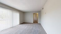 This single-floor, 2-bedroom, 2-bathroom unit offers a well-organized layout designed for comfortable everyday living. The living room is a generously sized, carpet-floored space featuring a large sliding glass door with vertical blinds that allows abundant natural light to pour in. Neutral gray walls and a textured ceiling carry throughout, creating a consistent, clean aesthetic. The kitchen is efficiently arranged in an L-shape, featuring warm oak-toned cabinetry, light countertops, and white appliances including a refrigerator, electric range, and dishwasher. A peninsula counter with a breakfast bar overhang opens to the adjacent dining or entry area, which features durable wood-look vinyl flooring. The primary bedroom offers a comfortable footprint with plush carpet flooring, a bifold-door closet with built-in rod and shelf, and direct access to one of the two bathrooms. The second bedroom is equally well-appointed with carpet flooring and a closet. The two bathrooms include one with a tiled tub-shower combo and wood-look vinyl flooring, and a second featuring a clean, all-white walk-in shower with tile surround. A connecting hallway provides smooth flow between the private and communal areas of the home.
