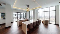 This elegant single-floor space is designed with a refined, contemporary aesthetic that seamlessly blends comfort and sophistication. Rich herringbone-patterned hardwood flooring flows throughout, anchoring each distinct zone and lending a warm, cohesive character to the open layout.

The expansive dining area is a standout feature, accommodating a long communal table surrounded by upholstered chairs, set atop a textured blue area rug. Above, warm wood ceiling beams add architectural depth, while floor-to-ceiling windows frame sweeping city views and flood the space with abundant natural light. Sliding glass doors provide direct access to an outdoor terrace.

The kitchen area is equally impressive, featuring a large white-topped island with bar seating, complemented by a sculptural gold pendant light fixture and a tray ceiling with warm ambient uplighting. Additional counter space and appliances make this a fully functional, high-end culinary environment, again framed by expansive windows overlooking the cityscape.

The lounge area provides a more intimate retreat, centered around a sleek fireplace set against a bold geometric-patterned accent wall in muted blue-grey tones. Exposed wood ceiling beams echo those in the dining area, while plush gold velvet sofas and tufted seating create an inviting atmosphere ideal for relaxed gatherings. A wall-mounted display screen adds a modern amenity to this comfortable social hub.