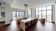 This elegant single-floor space is designed with a refined, contemporary aesthetic that seamlessly blends comfort and sophistication. Rich herringbone-patterned hardwood flooring flows throughout, anchoring each distinct zone and lending a warm, cohesive character to the open layout.

The expansive dining area is a standout feature, accommodating a long communal table surrounded by upholstered chairs, set atop a textured blue area rug. Above, warm wood ceiling beams add architectural depth, while floor-to-ceiling windows frame sweeping city views and flood the space with abundant natural light. Sliding glass doors provide direct access to an outdoor terrace.

The kitchen area is equally impressive, featuring a large white-topped island with bar seating, complemented by a sculptural gold pendant light fixture and a tray ceiling with warm ambient uplighting. Additional counter space and appliances make this a fully functional, high-end culinary environment, again framed by expansive windows overlooking the cityscape.

The lounge area provides a more intimate retreat, centered around a sleek fireplace set against a bold geometric-patterned accent wall in muted blue-grey tones. Exposed wood ceiling beams echo those in the dining area, while plush gold velvet sofas and tufted seating create an inviting atmosphere ideal for relaxed gatherings. A wall-mounted display screen adds a modern amenity to this comfortable social hub.