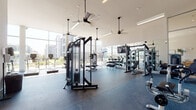 This contemporary fitness center is designed to support a full range of workout routines in a bright, open environment. The space is anchored by soaring ceilings and expansive floor-to-ceiling windows that flood the interior with natural light and provide views of the surrounding outdoor landscape. Durable rubber flooring covers the entire facility, offering both safety and comfort underfoot.

The main workout area is outfitted with a diverse selection of professional-grade equipment, including cable machines by TRUE Fitness, selectorized strength machines, adjustable benches, and a full rack of dumbbells spanning a wide weight range. Large wall-mounted mirrors line one side of the space, visually expanding the room and allowing users to monitor their form. A flat-screen television is mounted for added entertainment during workouts.

A dedicated functional training zone houses an impressive array of accessories, including kettlebells, medicine balls, battle ropes, a heavy bag, resistance bands, and plyo boxes, all neatly organized on storage racks. Cardio equipment, including treadmills and stationary bikes, is also available throughout the space.

Industrial-style ceiling fans hang from above, ensuring consistent airflow throughout the facility. The clean, modern aesthetic is completed by recessed and linear LED lighting, creating an energizing atmosphere suited for both focused training and casual exercise.
