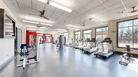 Located at 1111 W Stadium Dr in Fayetteville, Arkansas, this fitness center offers a generously sized, single-floor layout designed to accommodate a wide range of workout needs. The open floor plan is finished with durable, smooth flooring and features a drop-panel ceiling with multiple ceiling fans and recessed fluorescent lighting, ensuring a well-ventilated and bright environment throughout.

The cardio zone lines the windowed perimeter, where large framed windows flood the space with natural light and provide views of the outdoor surroundings. Treadmills, ellipticals, and stationary bikes are positioned to take full advantage of this natural light. A dedicated strength training area features a variety of cable machines and selectorized equipment, anchored by a bold red accent wall that adds a dynamic, energetic aesthetic to the space.

A separate section of the facility houses additional strength machines and a wall-mounted flat-screen TV, complemented by a rack of free weights and benches for versatile training options. Wall mirrors enhance the sense of depth and allow for proper form checks during exercise. Discreet doorways provide access to ancillary rooms, keeping the main workout floor unobstructed. The overall design is clean, functional, and purpose-built to support a comprehensive fitness experience.