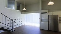 This two-story space offers a striking open-concept layout defined by soaring double-height ceilings that lend an expansive, airy quality to the main living area. Dark hardwood flooring runs throughout the lower level, providing a warm contrast to the crisp white walls and clean architectural lines. A large sliding glass door dressed with vertical blinds floods the space with natural light, creating a bright and welcoming atmosphere.

A distinctive white tiled accent wall adds textural interest and a mid-century modern aesthetic to the main living area. A pendant light fixture hangs gracefully from above, contributing to the overall ambiance. The upper level, accessible via a staircase visible from the main floor, features an open mezzanine-style overlook that connects the two floors visually, enhancing the sense of vertical space.

A convenient stacked washer and dryer unit is integrated directly into the main living area, offering in-unit laundry access. Structural columns and clean-edged walls define the spatial zones without closing off the open feel. Overall, the space combines functional living elements with architectural character, making it a practical yet visually dynamic environment.