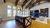 Located at 15155 Ollie Dr in Charlotte, NC, this single-floor community space is defined by its dramatic vaulted ceilings adorned with rich, dark-stained exposed wood beam trusses that immediately draw the eye upward and establish an inviting architectural character throughout. Warm hardwood flooring runs continuously across the open layout, unifying the distinct functional zones within the space.

The kitchen area is a standout feature, boasting a large central island with a crisp white countertop, dark wood paneling with decorative X-detail, and industrial-style bar seating. Flanking the island is a full run of dark cabinetry topped with light stone counters, complemented by a bold geometric tile accent wall in deep blue tones. Generous windows along the exterior walls flood the kitchen with abundant natural light and provide views of the surrounding community.

The living area offers a comfortable and sophisticated lounge environment centered around a sleek fireplace framed by built-in shelving units with fluted wood detailing, housing a wall-mounted television above. Plush, neutral-toned seating arrangements encourage relaxed gathering. A separate lounge nook features paired accent chairs, a round coffee table, bold botanical wall art, and access to private rooms enclosed with glass-panel sliding doors, ideal for smaller meetings or quiet retreats.