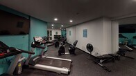 This compact yet fully outfitted fitness center is designed to meet a wide range of exercise needs within a single, efficiently organized space. The room features rubber flooring throughout, providing durability and safety underfoot during workouts. Recessed lighting illuminates the space from above, creating a functional and focused atmosphere.

The cardio area is anchored by multiple Spirit-brand treadmills lined along a teal accent wall, accompanied by a flat-screen television mounted at eye level for entertainment during workouts. A stationary recumbent bike and a rowing machine round out the cardio offerings. A standing fan provides additional air circulation throughout the space.

The strength training section houses a well-stocked dumbbell rack with a comprehensive range of free weights, a flat weight bench, and a selection of medicine balls and stability equipment neatly stored along the wall. Full-length mirrors are strategically positioned to allow users to monitor their form during exercise and visually expand the space.

A teal-painted entry door adds a pop of color that complements the accent wall, contributing to the room's cohesive design. An adjacent anteroom is accessible through the main gym door, offering additional utility space. Posted signage on the wall provides facility guidelines for users.