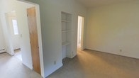This single-floor space features a straightforward and functional layout finished in neutral tones. The room is carpeted with beige wall-to-wall flooring that extends through the visible connected spaces, contributing to a cohesive and understated aesthetic. Walls are painted in a light gray-white hue, creating a clean, minimal backdrop throughout.

A notable built-in shelving unit is recessed into the wall, offering tiered open shelves above and a closed cabinet section below — providing practical storage without occupying additional floor space. This feature adds a degree of architectural interest to an otherwise simple interior.

A wood-toned door is visible to the left, likely leading to a closet or adjacent room, while an open doorway leads to a secondary space, suggesting a connected multi-room layout. The rooms benefit from natural light filtering through, keeping the interior bright and airy.

Overall, the space presents a clean, move-in ready environment well-suited for a variety of uses, with its neutral palette and built-in storage serving as practical, versatile foundations for personalization.