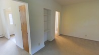 This single-floor space features a straightforward and functional layout finished in neutral tones. The room is carpeted with beige wall-to-wall flooring that extends through the visible connected spaces, contributing to a cohesive and understated aesthetic. Walls are painted in a light gray-white hue, creating a clean, minimal backdrop throughout.

A notable built-in shelving unit is recessed into the wall, offering tiered open shelves above and a closed cabinet section below — providing practical storage without occupying additional floor space. This feature adds a degree of architectural interest to an otherwise simple interior.

A wood-toned door is visible to the left, likely leading to a closet or adjacent room, while an open doorway leads to a secondary space, suggesting a connected multi-room layout. The rooms benefit from natural light filtering through, keeping the interior bright and airy.

Overall, the space presents a clean, move-in ready environment well-suited for a variety of uses, with its neutral palette and built-in storage serving as practical, versatile foundations for personalization.