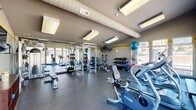 Located at 2880 Woodland Hills in Colorado Springs, CO, this fitness center is a comprehensively equipped exercise facility designed to support a full range of workout routines. The space is organized into distinct functional zones, making efficient use of its floor area.

The cardio section is positioned along a wall of large, floor-to-ceiling windows that flood the room with natural light and offer views of the surrounding outdoors. This area features multiple treadmills, an elliptical trainer, and a stationary cycle, each equipped with individual display monitors. The bright, airy atmosphere here creates an inviting environment for sustained cardio sessions.

The strength training area is anchored by a multi-station cable machine and a power rack, both positioned in front of large mirrors that visually expand the space and allow users to monitor their form. A dedicated free weights section includes a full rack of dumbbells and a variety of weighted medicine balls and stability balls. Flat and adjustable benches are available throughout.

The room features durable rubber flooring throughout, exposed HVAC ductwork along the ceiling, and bright overhead fluorescent lighting that ensures excellent visibility. A wall-mounted television provides entertainment. The neutral gray and yellow accent stripe wall color scheme gives the space a clean, functional aesthetic consistent with a professional fitness environment.