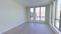 This single-floor space showcases a clean, contemporary design highlighted by light-toned hardwood-style flooring that runs throughout, creating a seamless and cohesive aesthetic. The interior is characterized by smooth white walls and crisp white ceilings, lending the space a fresh, minimalist atmosphere that maximizes the sense of openness.

A standout feature of the main living area is the pair of large, floor-to-ceiling windows framed in dark trim, which flood the interior with abundant natural light. These windows also offer direct views of the surrounding urban environment, including neighboring residential and commercial buildings as well as a city parking area, situating the space firmly within a vibrant metropolitan setting.

The layout is open and unencumbered, allowing for flexible furniture arrangement and making the space feel generously proportioned. Clean baseboards and subtle architectural details throughout reflect careful attention to finish quality. The overall ambiance is bright, airy, and modern — well-suited for those who appreciate understated, contemporary design with a strong connection to the energy of city living.