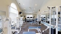 Located at 505 Regency Dr in Fayetteville, NC, this fitness center offers a thoughtfully designed, single-floor workout facility built to support a comprehensive exercise routine. The space is defined by soaring vaulted ceilings that lend an open, airy atmosphere, enhanced by recessed lighting and a ceiling fan that keep the environment comfortable during workouts. Large windows along the walls flood the interior with abundant natural light, creating an inviting and energizing ambiance. The flooring features durable, dark-toned athletic matting suited for heavy equipment and high-traffic use. The gym is well-stocked with a variety of strength training machines, including multi-station cable and weight systems by Paramount, as well as a selection of free-weight benches. Cardiovascular equipment, including treadmills and upright bikes, is positioned toward the rear of the space. A wall-mounted flat-screen television adds entertainment value for users during their sessions. Full-length mirrors line one wall, maximizing the sense of space while allowing users to monitor their form. The clean, neutral color palette with blue accent details throughout gives the facility a polished, professional appearance. Overall, this fitness center provides a well-rounded and welcoming environment for residents seeking a convenient on-site workout facility.