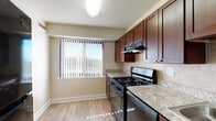 This single-floor residence offers a practical and comfortable layout across several interconnected rooms. The kitchen serves as a functional highlight, showcasing rich dark-wood cabinetry, light granite-style countertops, and a neutral tile backsplash. Black appliances, including a gas range with overhead ventilation and a dishwasher, are neatly integrated alongside a stainless steel sink. Light hardwood-style flooring and a window with vertical blinds bring brightness to the space.

The living areas feature plush grey carpeting and neutral beige walls, creating a calm, cohesive atmosphere throughout. Large windows dressed with vertical blinds allow generous natural light into each room, with views of surrounding greenery. A ceiling fan in the main living area adds both function and a finishing detail to the textured ceiling above.

The bathroom is notably well-finished, featuring floor-to-ceiling warm-toned stone tile surrounding a full soaking tub and shower combination. A wall-mounted mirror, a vanity with sink, and clean white fixtures complete the space with a polished appearance.

A utility closet with ventilated doors provides practical storage, while the overall floor plan, visible in the 3D model, reveals a logical and spacious arrangement of rooms connected by a central corridor.