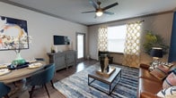 This single-floor space combines contemporary style with functional design, offering a welcoming and cohesive living environment. The main living area is a standout feature, showcasing rich hardwood-style flooring that runs throughout, complemented by neutral grey walls and crisp crown molding that add a polished, finished look to the space.

Natural light flows generously into the living room through multiple large windows dressed with bold gold and white patterned curtains, creating a bright and inviting atmosphere. A ceiling fan with an integrated light fixture is centrally positioned, providing both comfort and additional illumination.

The living area is thoughtfully laid out to accommodate both relaxation and casual dining. A rustic grey sideboard serves as a media console along one wall, while a modern rectangular coffee table anchors the seating area over a plush blue area rug. The adjacent dining nook features coordinating teal accent chairs, tying the color palette of the space together seamlessly.

A glass-paneled door offers access to an outdoor area, further enhancing the sense of openness and connectivity to the exterior. Overall, the space presents a stylish blend of warm textures, contemporary furnishings, and practical layout that makes it both comfortable and visually appealing.
