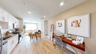 Located at 13611 Perry Rd in Houston, TX 77070, this single-floor residence offers a cohesive, open layout bathed in a warm, neutral palette throughout. Light wood-style flooring flows seamlessly across the entire living space, lending a sense of continuity and spaciousness.

The entry area transitions naturally into a versatile workspace featuring a dedicated desk area, framed artwork, and recessed lighting overhead. This flows into an open dining and living zone, where a ceiling fan with a light fixture crowns the living area, and large windows dressed with sheer curtains and horizontal blinds invite generous natural light. The living space accommodates both a comfortable seating arrangement and a defined dining area.

The kitchen is outfitted with white shaker cabinetry, granite countertops, a stainless steel appliance suite including a gas range, built-in microwave, refrigerator, and dishwasher, complemented by light wood flooring.

The bedroom features wide windows for ample natural light and access to a walk-in closet with multi-level hanging rods and shelving. The bathroom includes a granite-topped vanity with white cabinetry, a tub-shower combination with tile surround, and wood-style flooring throughout.
