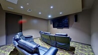 This single-floor media room is purpose-built for an immersive home theater experience. The room is finished with neutral taupe-toned walls and recessed LED ceiling lighting that creates a cinematic ambiance while maintaining functional visibility throughout the space. A large flat-screen television is prominently wall-mounted at the focal point of the room, accompanied by a soundbar and flanking wall-mounted surround sound speakers, delivering a comprehensive audio-visual setup.

Seating consists of multiple blue leather reclining chairs and sofas with white accent stitching, arranged in a curved, theater-style configuration to optimize sightlines to the screen. The recliners feature built-in cupholders, adding to the comfort and functionality of the space. The flooring is covered in a bold patterned carpet with a geometric design in brown and lime green tones, adding a striking visual contrast to the darker walls and furniture.

A single door provides access to the adjoining area, reinforcing the room's enclosed, dedicated design. The combination of controlled lighting, quality audiovisual equipment, and plush reclining seating makes this media room a well-appointed, functional entertainment space.