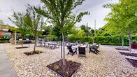 Located at 3201 First Ave in Columbus, Georgia, this expansive outdoor space offers a thoughtfully designed environment for gathering and recreation. The grounds are finished with a gravel and pea stone surface, providing a clean, low-maintenance aesthetic that complements the natural surroundings. Numerous maturing trees are planted throughout the space in mulched beds, offering shade and a sense of greenery that softens the open-air setting. String lights are strung across the area on tall poles, creating a warm and inviting ambiance suited for both daytime and evening use. Seating is varied and plentiful, including multiple picnic tables for group dining, clusters of Adirondack chairs arranged around a central fire pit for relaxed conversation, and hanging egg chairs that add a casual, modern touch. A covered pergola structure anchors one section of the space, providing a shaded retreat and additional seating beneath its canopy. Paved pathways wind through the gravel areas, offering easy circulation across the grounds. Lush green hedgerows and mature trees line the perimeter, creating a natural boundary that lends privacy. The backdrop includes views of neighboring brick architecture, situating this outdoor venue within the broader urban character of Columbus.