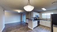 This single-floor space showcases an open-concept layout that integrates the kitchen and living area into one cohesive, functional environment. The kitchen is outfitted with crisp white shaker-style cabinetry, offering ample storage both above and below the countertops. Dark stone countertops provide a striking contrast against the white cabinets and are carried across both the perimeter counters and a peninsula, which serves as a natural divider between the kitchen and living area. Stainless steel appliances, including a range with electric cooktop and an over-the-range microwave, complete the modern kitchen aesthetic.

A pendant light with a frosted glass shade hangs prominently over the peninsula, adding a focal point while providing functional task lighting. The textured ceiling features additional flush-mount lighting throughout. Wide-plank wood-look flooring runs continuously across both the kitchen and living area, reinforcing the open, unified feel of the space. Neutral gray walls create a versatile backdrop. Sliding glass doors at the far end of the living area invite natural light and provide access to an outdoor area.