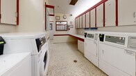Located at 345 Sentinel, CA, this shared laundry facility is a single-floor space designed for practical, efficient use. The room is finished with a distinctive cream and red color scheme, with bold red trim accenting the walls, cabinetry, and countertops, creating a cohesive and easy-to-maintain aesthetic throughout.

The main laundry area is equipped with multiple coin-operated washers and dryers lined along one wall, accompanied by an extensive run of upper wall-mounted cabinets that provide ample storage above the machines. A continuous countertop with a built-in utility sink runs along the adjacent wall, offering a dedicated space for folding, sorting, and hand-washing. Additional upper cabinets extend above the counter, maximizing storage capacity.

A separate alcove area features a bank of individual storage lockers, neatly arranged in a grid formation, providing secure personal storage for users. Posted signage and operating instructions are prominently displayed throughout the room for ease of use.

The flooring throughout is a speckled terrazzo-style material, durable and easy to clean, well-suited to the functional nature of the space. Overhead ventilation and recessed lighting ensure adequate airflow and illumination. The layout is straightforward and practical, making this facility a reliable and organized shared amenity.