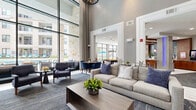 Located at 770 N Eldridge Pkwy in Houston, TX, this thoughtfully designed communal space offers a sophisticated blend of modern architecture and inviting comfort. The living area commands attention with its dramatic double-height ceilings, floor-to-ceiling windows, and an abundance of natural light that floods the interior. Textured accent walls — including mosaic tile columns and a sculptural wave-panel feature wall — add visual depth and elegance throughout. The lounge is anchored by a plush neutral sofa, tufted armchairs, and a live-edge wood dining table positioned beside expansive windows overlooking a lush courtyard with a pool and outdoor seating. The kitchen and dining area is appointed with rich wood cabinetry, granite countertops, exposed wood beam ceilings, and slate-blue subway tile backsplash, creating a warm yet contemporary atmosphere. Pendant lighting, marble-look tile flooring, and a built-in media credenza further enhance the refined aesthetic. This space seamlessly connects its lounge, dining, and kitchen zones, offering a cohesive and functional environment well-suited for gathering and community engagement.