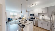 This thoughtfully designed single-floor residence offers a seamless blend of modern aesthetics and functional living. The open-concept layout connects the kitchen and living area, creating an inviting, airy atmosphere filled with natural light from large windows that frame outdoor views. The kitchen is a standout feature, showcasing gray shaker-style cabinetry with matte black hardware, granite countertops, a central island with bar seating, and a full suite of stainless steel appliances including a refrigerator, range, microwave, and dishwasher. Pendant lighting above the island adds a refined touch, while light wood-tone flooring flows throughout the main living areas. The living room is bright and welcoming, featuring a generous window with outdoor views and ample space for comfortable seating arrangements. The bedroom continues the clean, contemporary feel with plush carpeting, a large window overlooking the exterior, and warm, neutral tones that create a restful environment. The bathroom is finished with marble-style tile flooring, a granite-topped vanity, and a full soaking tub with a tiled surround. A smart-lock entry door completes this well-appointed residence.
