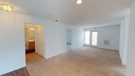 This single-floor space presents a clean, functional layout centered around a generously sized open living area finished with wall-to-wall beige carpeting that adds warmth underfoot. The room is bathed in natural light courtesy of a set of French doors along the far wall, which also offer potential outdoor access. Recessed ceiling lighting supplements the natural light, ensuring the space remains well-lit throughout the day. The white walls and white-trimmed doorways lend a bright, neutral aesthetic throughout.

Adjacent to the main living area, an open doorway leads to a secondary room, also finished with carpeting, providing flexible use as a bedroom or additional living space. To the left, a bathroom is visible and features warm wood cabinetry and vanity lighting above the mirror, offering a practical and functional layout. A wall-mounted HVAC vent is positioned near the French doors, ensuring climate control throughout the space.

Overall, the space maintains a straightforward, move-in-ready character with neutral finishes, practical connectivity between rooms, and ample natural light.