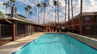 This single-level outdoor amenity space is a standout feature of a brick-exterior residential community, offering a well-designed environment for recreation and relaxation. The centerpiece is a generously sized swimming pool with crystal-clear blue water, surrounded by a spacious concrete pool deck ideal for lounging. The pool area is enclosed by a secure wrought-iron-style fence and is framed by a striking row of tall palm trees that reach toward a bright, open sky, creating a resort-like atmosphere.

Adjacent to the pool is a covered pavilion area with a wood-paneled ceiling and open-air views of the surrounding courtyard. This sheltered space connects to a raised wood deck supported by sturdy columns, offering a comfortable shaded area for gathering. The deck overlooks the pool and the broader community grounds, which feature brick buildings with pitched rooflines.

A covered walkway runs along the side of the building, lined with a series of white-framed French doors that allow natural light to filter through. This corridor also features a vending machine and fire safety equipment, adding practical functionality to the space. The combination of thoughtful architectural details, lush landscaping, and well-maintained amenities makes this outdoor area both inviting and highly functional for residents.