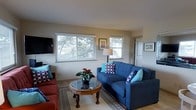 This single-floor space centers around a well-appointed living area designed for comfort and casual elegance. The room is filled with natural light streaming through multiple white-framed windows that offer pleasant views of mature trees and greenery outside, creating a warm and inviting atmosphere. The neutral, light-toned walls provide a versatile backdrop that complements the carefully chosen furnishings. A navy blue sofa, accented with teal and patterned throw pillows, serves as the focal point of the seating arrangement, paired with a rich red sofa that adds a bold contrast. A ornate wooden coffee table with a glass top anchors the seating area, styled with a decorative plant centerpiece. A patterned area rug defines the space and adds visual texture underfoot. A wall-mounted flat-screen television is positioned for easy viewing, while framed artwork on the walls adds a personal touch. The entry door is visible to one side, suggesting a practical and open layout. Overall, the space exudes a relaxed, homey ambiance with a thoughtful mix of color, texture, and natural light.
