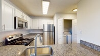 Located at 6430 Brookstone Ln in Fayetteville, NC, this single-story unit offers a practical and comfortable layout with thoughtfully selected finishes throughout. The kitchen serves as a functional centerpiece, showcasing granite countertops, white cabinetry with brushed nickel hardware, and a full suite of stainless steel appliances including an electric range, over-the-range microwave, and refrigerator. A deep stainless steel sink with a modern pull-down faucet adds convenience, and a dedicated in-unit laundry closet is conveniently accessible directly from the kitchen area.

The living areas feature light-toned hardwood-style flooring, neutral gray walls, and a ceiling fan with integrated lighting, creating a bright and open atmosphere. A window allows natural light to filter into the space. Bedroom areas are finished with plush wall-to-wall carpeting, providing a softer, more comfortable feel, and feature ample wall space and closet access.

The bathroom is clean and well-finished, featuring a white vanity with granite countertops, a large mirror, stylish globe lighting fixtures, and wood-style flooring that ties the space together cohesively. Throughout, textured ceilings and consistent neutral tones contribute to a tidy, move-in-ready ambiance.