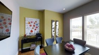 This single-floor space showcases a thoughtfully designed dining area that serves as its visual centerpiece. The room is defined by striking yellow-green accent walls that create a warm, energetic atmosphere, contrasting with neutral gray-toned walls and a crisp white ceiling. Recessed lighting complements the natural light that filters in through a large window fitted with horizontal blinds, offering views to the outdoors.

The dining area features a dark-toned wood table surrounded by contemporary gray upholstered chairs, providing comfortable and stylish seating. A decorative wire bowl with colorful yarn balls serves as a charming table centerpiece. Along one wall, a low-profile black media console holds a printer and is adorned with a bold abstract canvas artwork featuring colorful spheres, adding an artistic focal point to the room. A pair of narrow vertical mirrors on an adjacent wall enhances depth and reflects light throughout the space.

The hardwood flooring in a warm honey tone runs throughout, grounding the room with a natural, inviting quality. The overall ambiance is modern and vibrant, blending functional furnishings with expressive design elements.