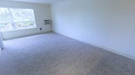 This single-floor space offers a clean, straightforward layout with a neutral, functional design throughout. The main living area is finished with wall-to-wall carpeting in a light beige tone, providing a soft and comfortable underfoot feel. The walls are painted in a crisp, off-white shade that helps maximize the sense of brightness and openness in the room. A window along one wall allows natural light to filter into the space, contributing to an airy atmosphere. A wall-mounted window air conditioning unit provides climate control, accompanied by a floor-level heating vent nearby, ensuring year-round comfort. The baseboards are clean and white, complementing the overall neutral palette of the interior. The room is open and unobstructed, offering a blank canvas that can be configured to suit a variety of uses and furniture arrangements. The overall ambiance is simple and practical, with an emphasis on functionality. The space is well-suited for those looking for a move-in ready environment with essential utilities already in place and the flexibility to personalize the interior to their own taste and needs.