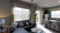 This single-story space centers around a well-appointed living area that showcases a contemporary design aesthetic. The room is anchored by a large dark sectional sofa adorned with patterned accent pillows, positioned to take full advantage of the abundant natural light streaming through multiple large windows dressed with layered blinds and decorative patterned drapes. A sliding glass door provides direct outdoor access while further brightening the space.

The living area features smooth, light-toned walls complemented by dark hardwood-style flooring that runs throughout, creating a clean contrast. A ceiling fan is centrally positioned overhead, adding both function and a finishing touch to the room. Decorative elements include a tall indoor plant, a metallic coffee table, and a console table styled with bottle arrangements, contributing to the curated, modern feel.

A hallway leading into the living room is adorned with sunburst wall art and teal canvas paintings, adding character and visual interest. The kitchen is partially visible from the living area, featuring stainless steel appliances and a light-toned countertop with bar seating, reinforcing the open, connected layout of the space.
