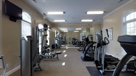 This fitness center offers a comprehensive workout environment designed to accommodate a variety of exercise routines. The single-floor space is thoughtfully laid out, with cardio machines — including treadmills and an elliptical — lining one side of the room, while a dedicated strength training area featuring cable machines, free weights, and a weight bench occupies the other. Large floor-to-ceiling mirrors span the length of one wall, enhancing the sense of spaciousness and allowing users to monitor their form during workouts. The interior is finished with warm, neutral-toned walls and elegant crown molding, lending the space a polished, upscale feel. Bright fluorescent panel lighting ensures the entire room is well-illuminated throughout the day. An arched window along the far wall invites natural light into the space, adding an airy quality to the environment. The flooring transitions between a smooth, easy-to-clean surface in the cardio zone and carpet in the strength area, providing both functionality and comfort. A wall clock and posted informational signage add practical touches to the well-organized layout. Overall, this fitness center strikes a balance between functionality and refined design.
